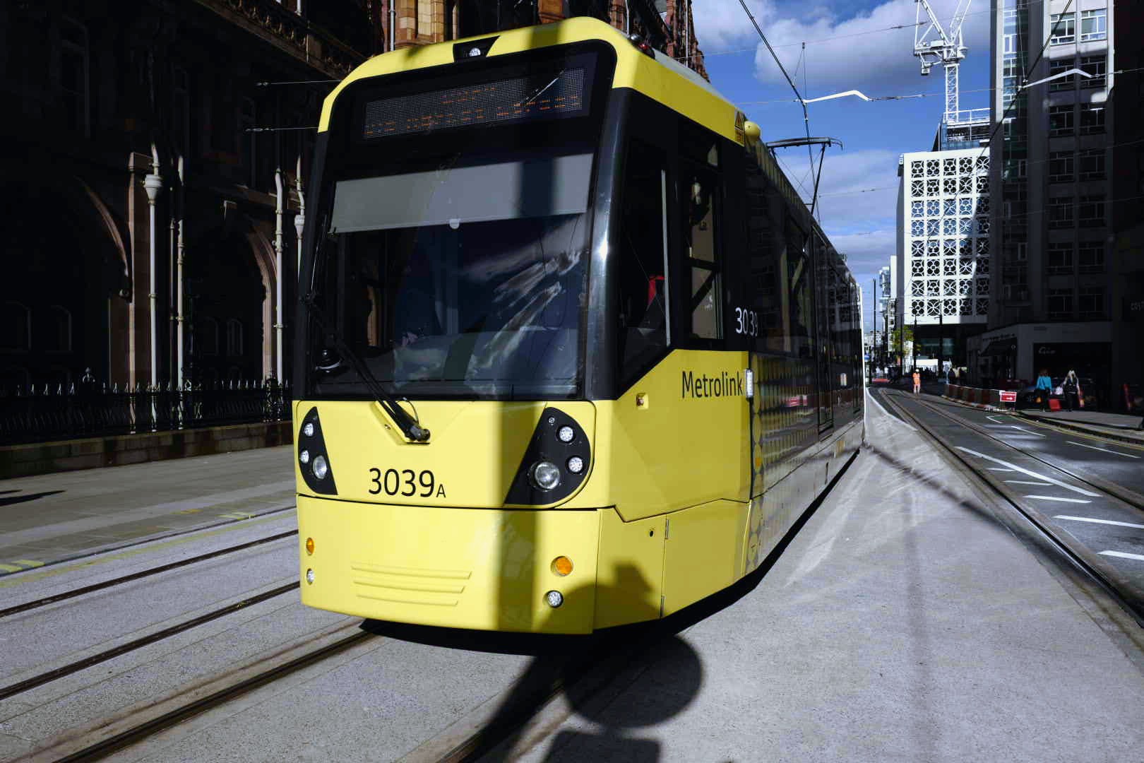 A yellow Manchester Metrolink tram on street-level tracks, with city-centre buildings behind.